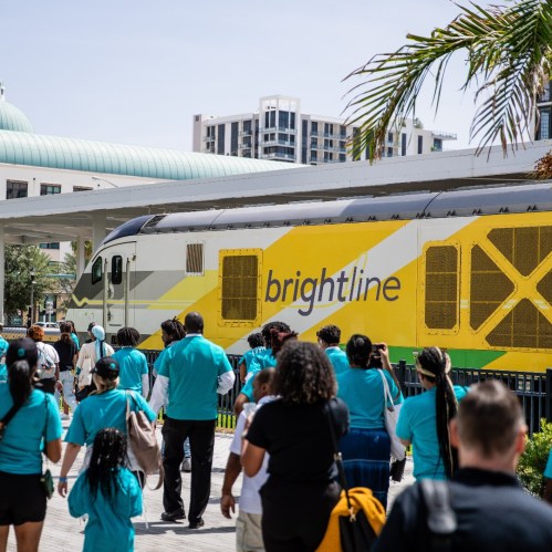 A Brightline train at a station with passengers in turquoise shirts waiting nearby under a sunny sky.
