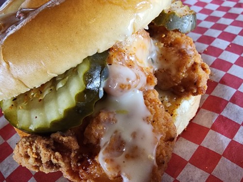 Close-up of a fried chicken sandwich with pickles and creamy sauce, served on a toasted bun with a checkered background.