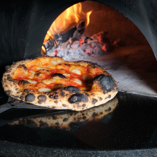 A freshly baked pizza with a charred crust being pulled from a wood-fired oven.