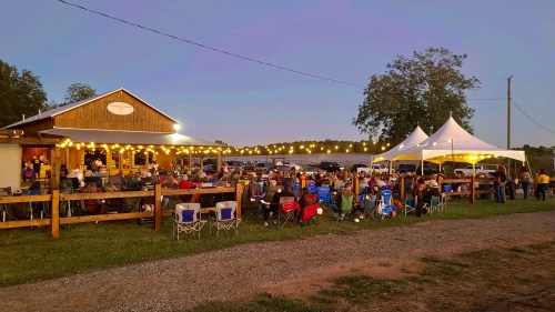 A lively outdoor gathering at dusk, with people seated at tables and a tent, surrounded by string lights and nature.