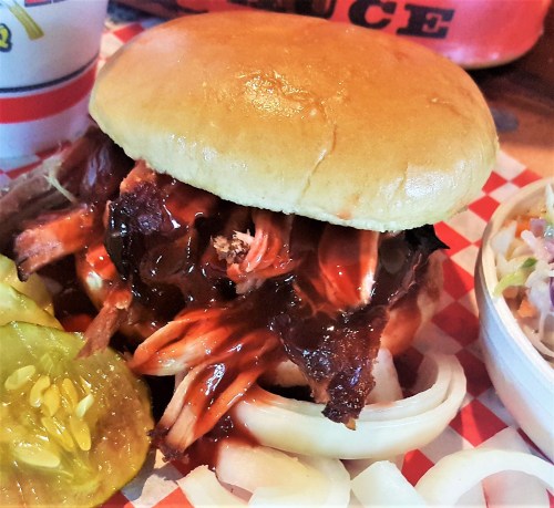 A close-up of a pulled pork sandwich with barbecue sauce, served with pickles and onions on a checkered plate.