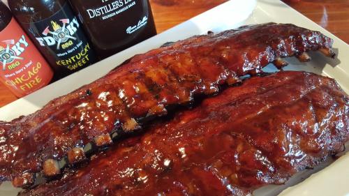 A plate of glazed barbecue ribs with bottles of sauce in the background.