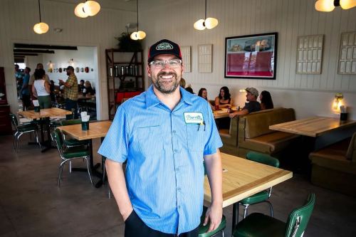 A smiling man in a blue shirt and cap stands in a bright restaurant with wooden tables and green chairs.
