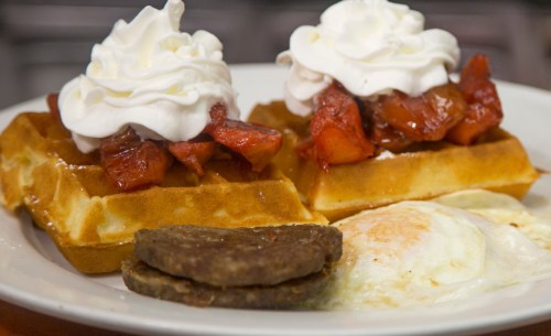 A plate of waffles topped with whipped cream and strawberries, served with two sausage patties and a fried egg.