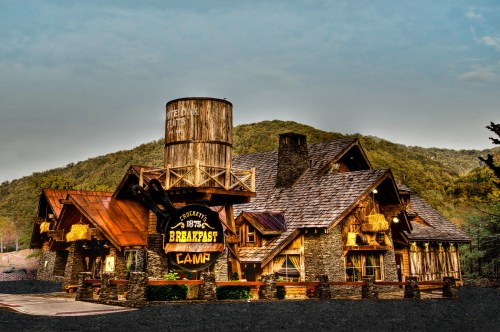 A rustic lodge-style building with a water tower, surrounded by mountains, featuring a sign for "Breakfast Camp."