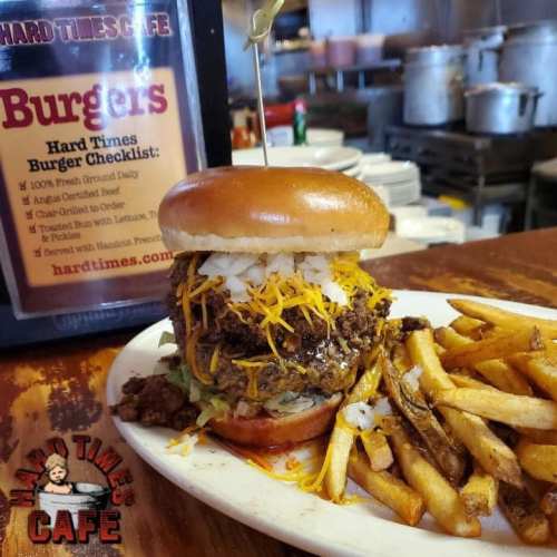 A towering burger with cheese, onions, and toppings, served with fries, next to a menu sign for Hard Times Cafe.
