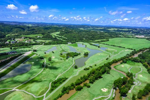 Aerial view of a lush green golf course with ponds and winding rivers, surrounded by rolling hills and blue skies.