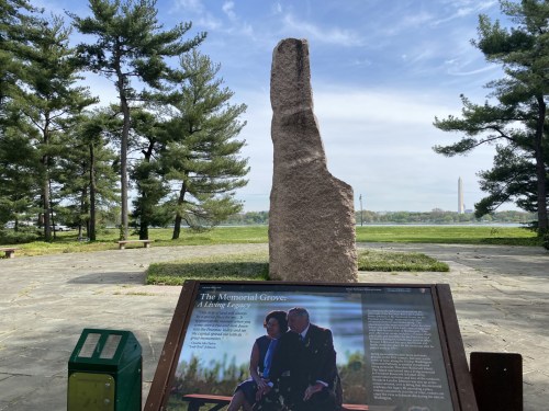 A stone monument in a park with a plaque, overlooking a body of water and trees, with distant structures visible.