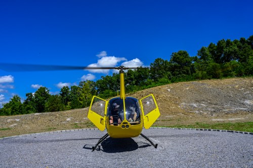 A yellow helicopter with open doors sits on a gravel pad under a blue sky with scattered clouds and green trees in the background.