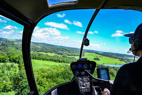 Aerial view from a helicopter cockpit, showcasing lush green landscapes and a blue sky with scattered clouds.