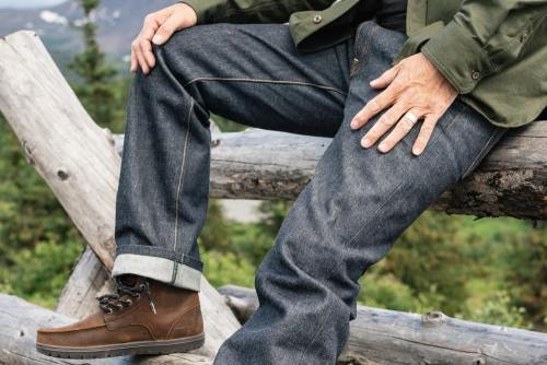 A person sits on a log, wearing dark jeans and brown boots, with a green jacket, surrounded by nature.