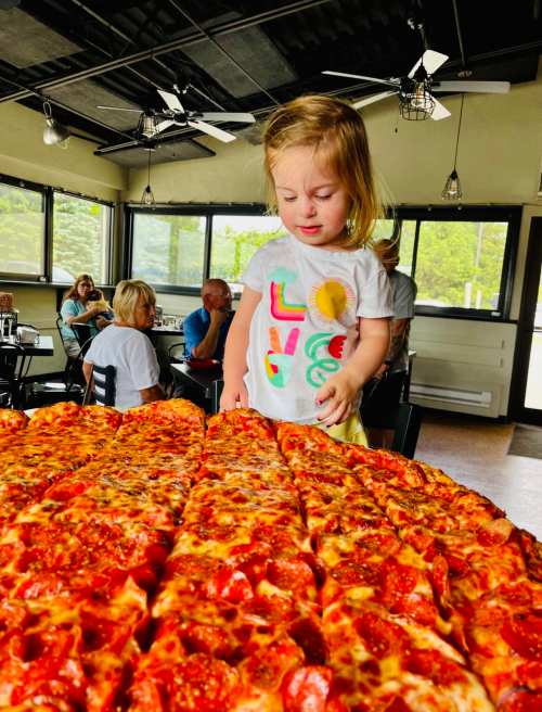 A young child stands in front of a large pizza in a restaurant, looking down at the food with curiosity.