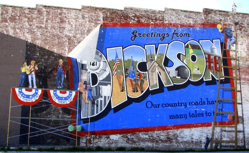 Mural reading "Greetings from Jackson" with colorful artwork and people painting on a ladder.