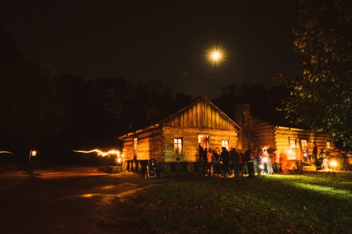A rustic cabin at night, illuminated by moonlight, with a group of people gathered outside. Stars twinkle above.