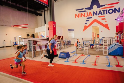Two children run on a red track in a colorful indoor obstacle course at Ninja Nation, with various equipment in the background.