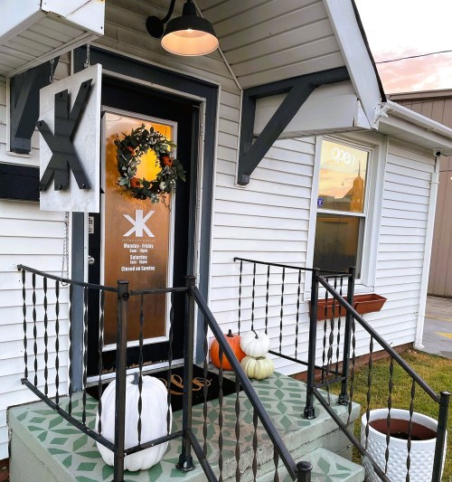 A cozy entrance with a wreath, pumpkins, and a decorative porch leading to a white house with a sign.