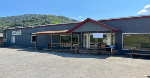 A gray building with a red roof and wooden porch, surrounded by greenery and mountains in the background.