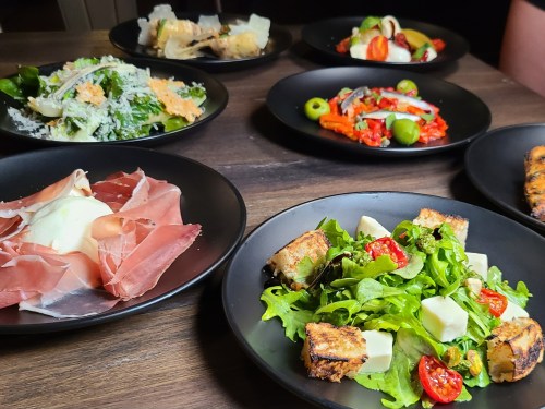 A variety of dishes on black plates, including salads, cured meats, and colorful vegetables, arranged on a wooden table.