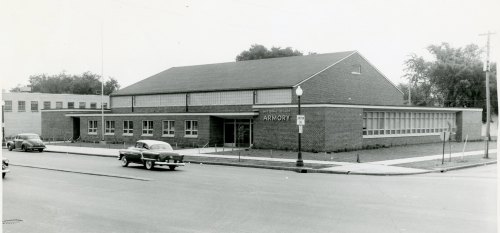 Black and white photo of a brick armory building with large windows, parked cars, and a tree-lined street.