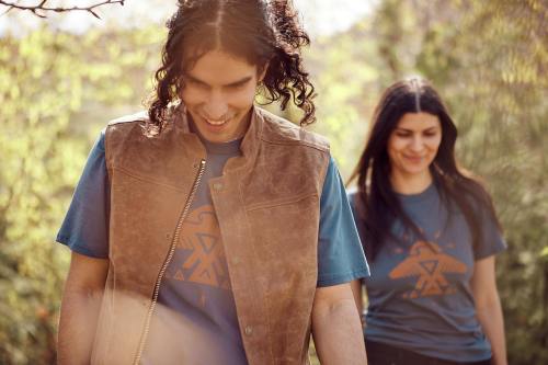 Two people outdoors, smiling and wearing matching blue t-shirts with a graphic design, surrounded by greenery.