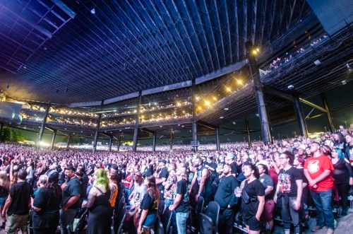 A large crowd of concertgoers fills an outdoor venue, illuminated by stage lights and surrounded by a modern structure.