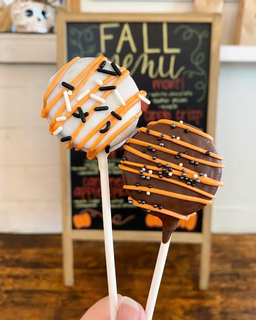 Two decorated cake pops on sticks, one white with orange and black drizzle, the other brown with orange stripes and sprinkles.