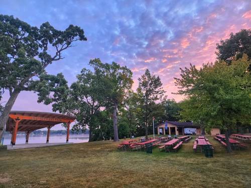 A scenic park at sunset with picnic tables, a pavilion, and trees by a lake, featuring colorful clouds in the sky.