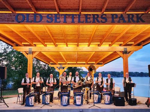 A brass band performs on stage at Old Settlers Park, with a scenic lake view in the background.