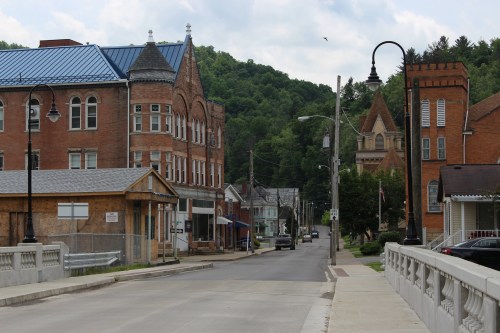 A quiet street in a small town, featuring historic brick buildings and green hills in the background under a cloudy sky.