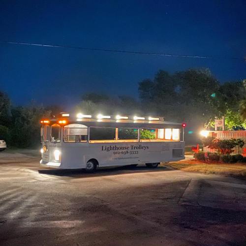 A brightly lit trolley bus parked at night, surrounded by trees and a quiet road.