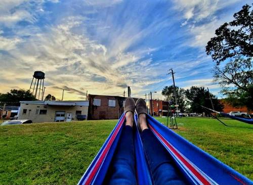 Person relaxing in a hammock with feet visible, surrounded by grass and a scenic sky with clouds.