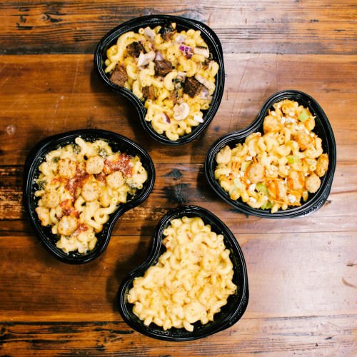 Four heart-shaped containers of macaroni and cheese with various toppings on a wooden table.