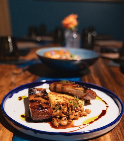 A plate featuring grilled ribs, black-eyed peas, and a slice of cornbread, with a blurred background of a dining setting.