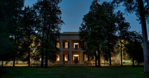 A grand, illuminated mansion surrounded by tall trees at dusk, showcasing classic architecture and a serene atmosphere.