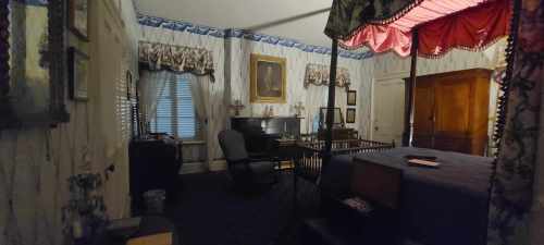 A vintage bedroom with a four-poster bed, desk, and period decor, featuring patterned wallpaper and a large window.