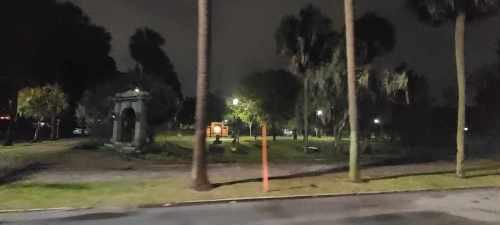 A dimly lit park at night with palm trees, a stone structure, and scattered lights in the background.