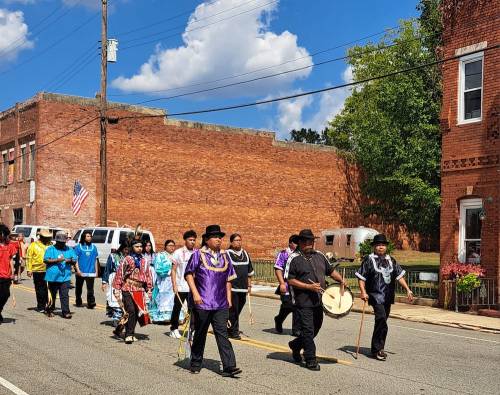 A group of people in colorful traditional attire march down a street, some playing drums, under a blue sky.