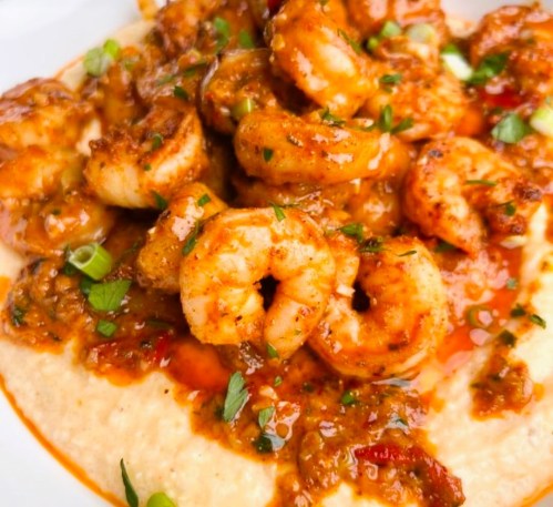 A close-up of shrimp in a spicy sauce served over creamy grits, garnished with green onions and herbs.
