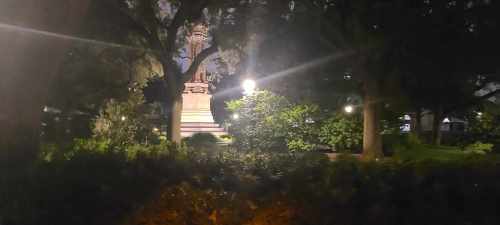 A dimly lit park scene at night featuring a statue surrounded by trees and greenery.