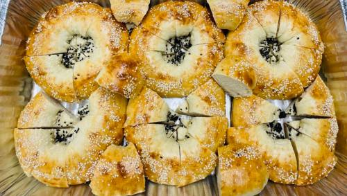 A tray of freshly baked bagels, some sliced, topped with sesame seeds and black seeds.