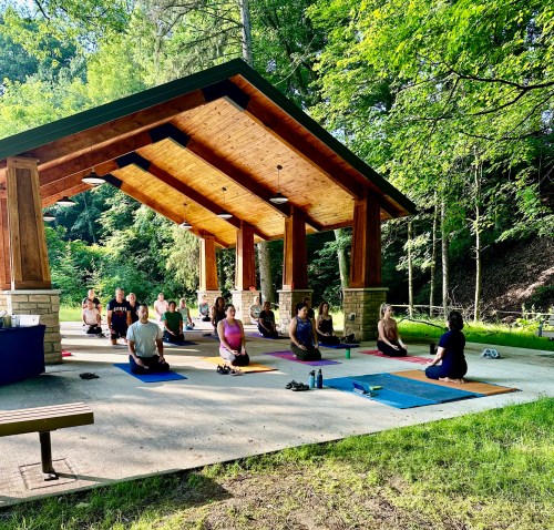 A group of people practicing yoga outdoors under a wooden pavilion surrounded by trees.