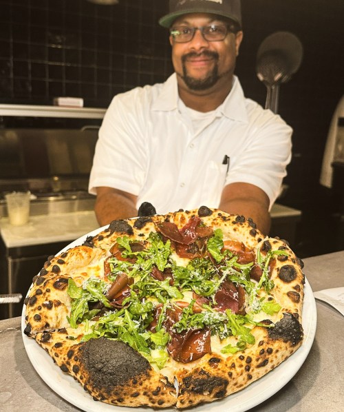 A chef proudly holds a freshly made pizza topped with greens and meats, showcasing its crispy crust in a kitchen setting.