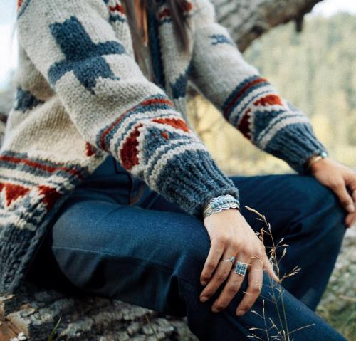 A person in a patterned sweater and jeans sits outdoors, resting their hand on their knee, surrounded by nature.