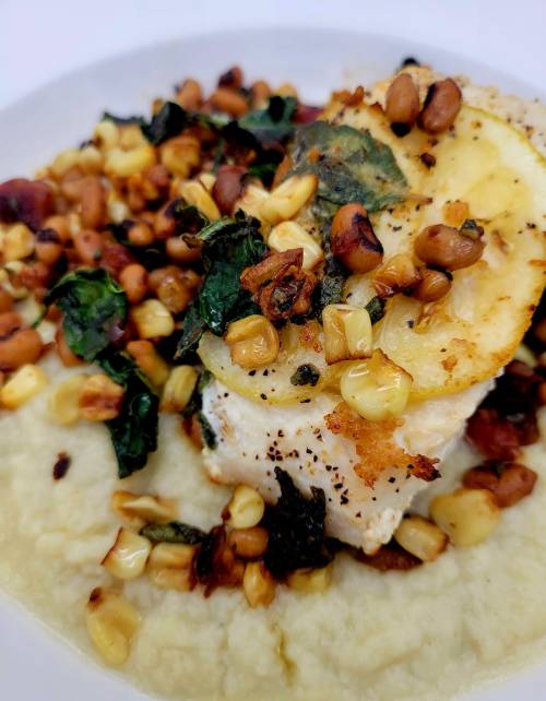 A plate of fish topped with sautéed greens, corn, and beans, served over a creamy base.