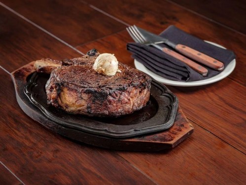 A juicy ribeye steak on a sizzling platter, topped with a dollop of butter, alongside utensils and a napkin.