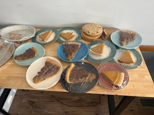A table with various slices of pie and cookies on plates, some covered with plastic wrap.