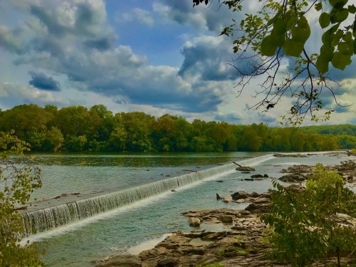 A serene river scene with a waterfall, surrounded by lush green trees and a cloudy blue sky.