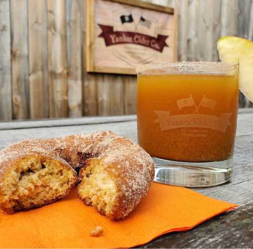 A glass of cider with a slice of lemon beside a cinnamon sugar donut on a wooden table.