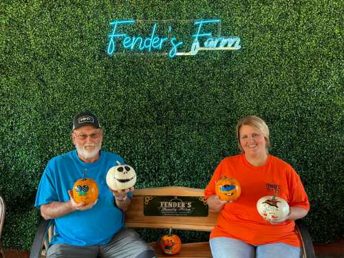 Two people sit on a bench at Fender's Farm, holding decorated pumpkins against a green backdrop with a neon sign.