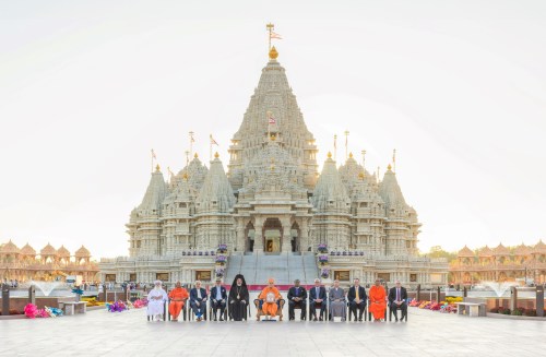 A group of people seated in front of a grand temple with intricate architecture, surrounded by flowers and a clear sky.